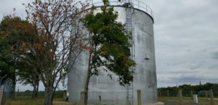 Ein großer, grauer Wasserspeicher mittem im grünen umgeben von ein paar Bäumen.