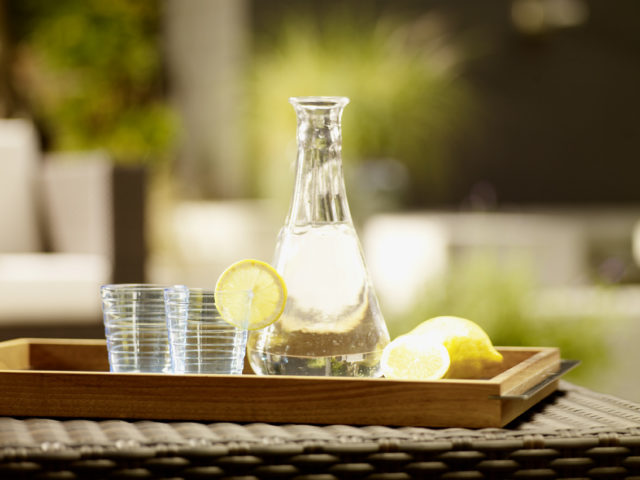 Eine Glaskaraffe gefüllt mit Wasser, daneben zwei leere Trinkgläser. Daneben liegt eine ganze Zitrone sowie einzelne Zitronenscheiben.
