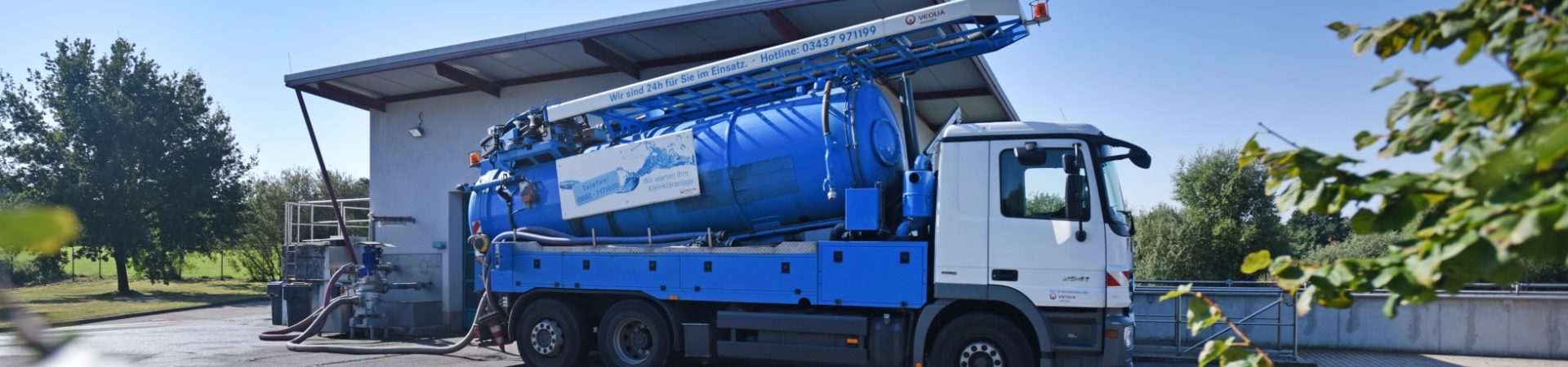 Ein blaues Kanalreinigungsfahrzeug steht am Straßenrand. Der große Tankwagen hat einen ausgefahrenen Saugschlauch, der in einen Kanalschacht führt. Das Fahrzeug ist dabei, Abwasser oder Schmutz aus dem Kanalsystem abzupumpen.