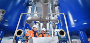 Innenansicht eines Wasserwerks: große blaue Behälter mit vielen Rohren. Zwei Mitarbeiter von Veolia haben einen großen Plan in der Hand.