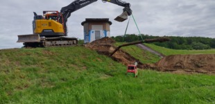 Ein gelber Bagger steht auf einem grünen Hügel inmitten einer ländlichen Umgebung. Der Bagger ist bei Erdarbeiten für die Installation neuer Wasserleitungen im Einsatz. Im Vordergrund ist aufgewühlte Erde zu sehen, die auf die laufenden Bauarbeiten hinweist.