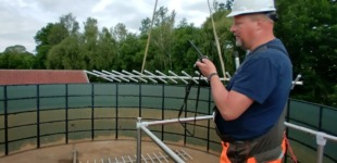 Ein Bauarbeiter steht auf einem Metallgerüst, das in einem leeren Abwasserbecken errichtet wurde. Das große Becken ist komplett entleert und zeigt deutliche Spuren von Abnutzung an den Betonwänden. Der Arbeiter, der sich etwa auf halber Höhe des Beckens befindet, führt Sanierungsarbeiten durch.