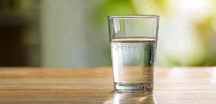 Ein klares Glas mit frischem Wasser steht auf einer hölzernen Oberfläche. Das Glas ist zur Hälfte gefüllt und kleine Luftbläschen haften an der Innenseite. Der Hintergrund ist unscharf und in warmen, grün-gelben Tönen gehalten, was dem Bild eine natürliche, frische Atmosphäre verleiht.